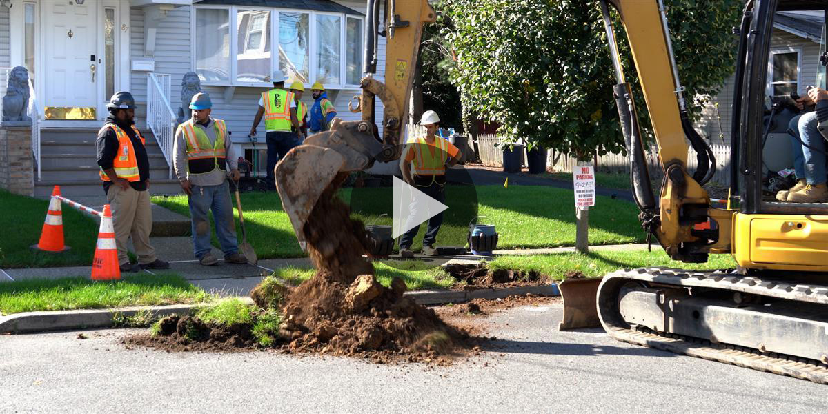 Construction crew operates and oversees excavator digging up street to reveal pipes 